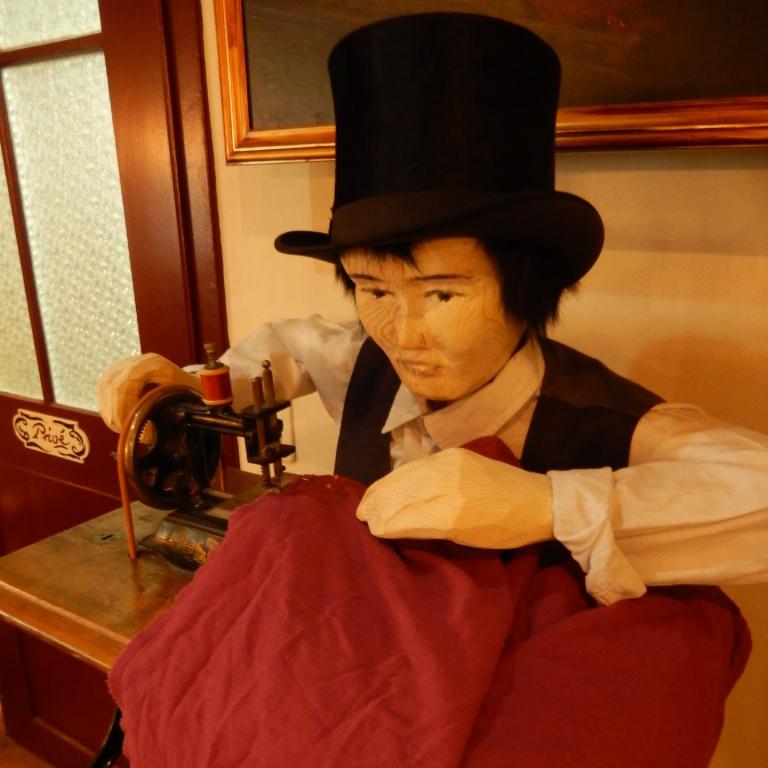 Mechanische Holzpuppe in einem schwarzen Top Hat und weißem Outfit sitzt bedeckt mit einem roten Tuch, neben einer antiken Nähmaschine.