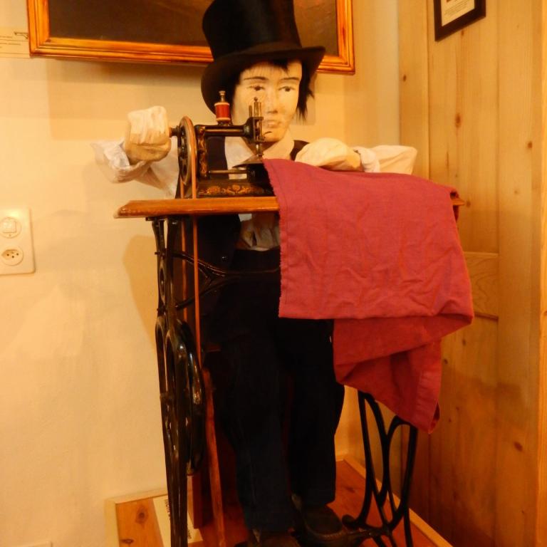 Eine mechanische Holzpuppe in Hut und Anzug, das in einer historischen Ausstellung mit roten Stoffen und einem Gemälde zu sehen ist.